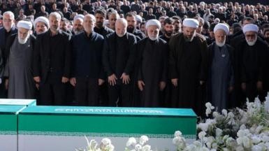 Iranian President Masoud Pezeshkian, Iran's Parliament speaker Mohammad Baqer Qalibaf and Chief Justice of Iran Gholam-hossein Mohseni Ejei with officials attend a prayer during a public farewell ceremony to pay their respects to late Supreme Leader Ayatollah Ali Khamenei. A large crowd can be seen in the background as everyone stands looking at the coffin