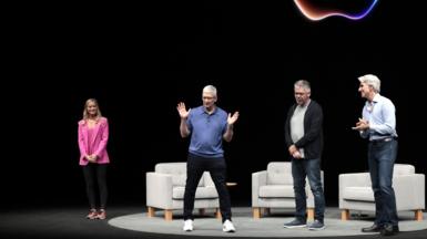 Apple CEO Tim Cook on a dark stage with a neon Apple logo behind him. He is wearing a blue polo sweater, black jeans and white sneakers and gesturing his hands, while three other people on stage look at him.