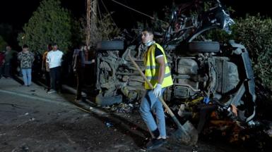 A young man in hi-vis holds a shovel in front of a car, on its side and destroyed by an apparent explosion. Other young men look on. It's dark and the car is on a street in front of a hedge.