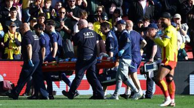 Footballer Conor Coady being carried off a football pitch on a stretcher by a team of male medics wearing dark colours while a player in a yellow shirt with red trim, red shorts, white socks and white boots is visible in the foreground and fans applauding are visible in the background