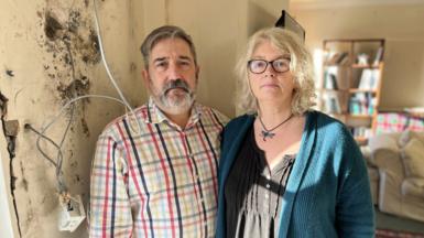 Tony and Bec Wadley standing next to a wall covered in black mould with electrical wires and sockets hanging from it