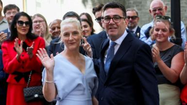 Andy Burnham stands next to his wife Marie-France Van Heel who is waving with crowds clapping the in the background