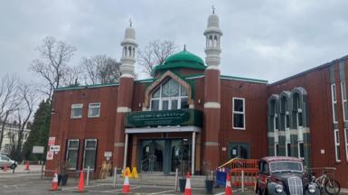 The entrance to a mosque, a brick building with dark green signage. The is a green dome shaped structure on the roof and two minaret towers with Islamic crescent moon sculptures on top. 
