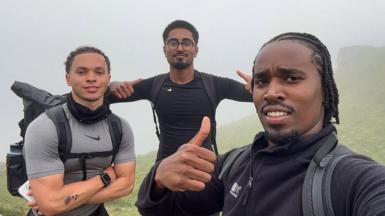 A selfie of three men standing on a foggy mountain. They all wear dark black, and grey clothing. Two men have their thumbs up while one stands with his arms folded. 