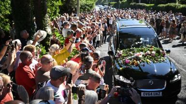 Bystanders flock towards a black hearse and throw flowers onto its bonnet as it drives through the town carrying the coffin of Bonnie Tyler.