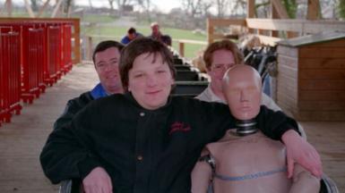 Justin Garvanovic sat in the front row of the Megafobia ride at Oakwood with his arm around a test dummy figurine. There are two men in the row behind. In the background more people are in the carts at the tail end of the rollercoaster with the green setting of Oakwood featuring trees and fields behind them.