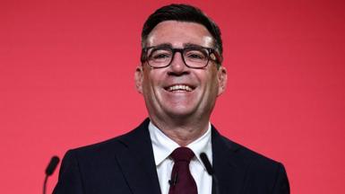 Andy Burnham in a dark suit in front of a red background