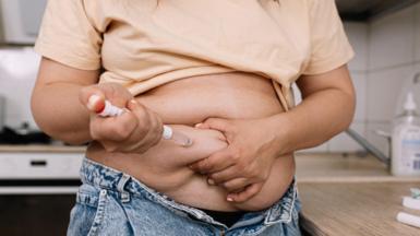 A woman uses a pre-filled injection pen to administer a dose. She is lifting her t-shirt to be able to inject.