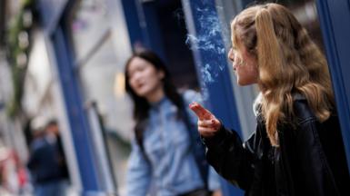 A young woman smokes outside a shop. She has long blonde hair and wears a leather jacket