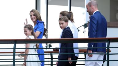 The Princess of Wales, Princess Charlotte, Prince George, and the Prince of Wales walk across a bridge at Wimbledon. Prince George has blonde hair and is wearing a navy suit. The Prince of Wales is  wearing a navy blazer and white trousers. The Princess of Wales has long brown hair and is wearing a blue dress. She is waving. Princess Charlotte has long brown hair and is wearing a white dress.