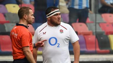 Referee Angus Gardner (left), wearing an orange shirt points his left index finger while he talks to England captain Jamie George, wearing all white with white and black tape around his head, with both looking to the right