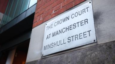 A close-up photograph of a metallic sign, secured to a brick wall, which reads 'The Crown Court at Manchester Minshull Street' in engraved lettering. 