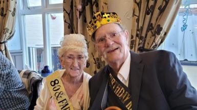 Ronald Parkin and his wife Patricia are sitting together and smiling at the camera. They are both wearing crowns and sashes across their chest to signify it's their birthday. Ronald is wearing a black suit jacket and white shirt and wears glasses. Patricia is wearing a cream cardigan and is also wearing glasses and gold hoop ear rings. 