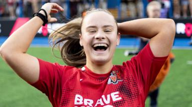 Ffion smiling on pitch in her red rugby kit. She is putting her hair in a ponytail while looking at the camera. 