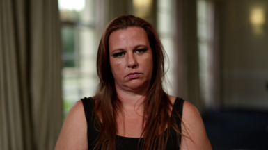 A woman wearing a black tank top and long reddish brown hair sits and stares at the camera.