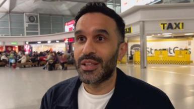 A male passenger with a white shirt and blue jacket looks adjacent to the camera with people sitting down next to a taxi area in Stansted Airport