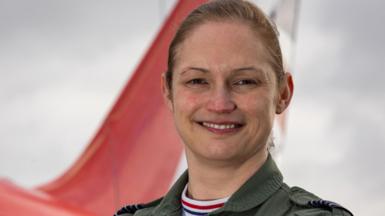 Head and shoulders shot of a smiling Wing Cdr Sasha Nash. She has fair hair which is tied back and is wearing overalls. There is the tail section of a Red Arrows jet in the background.