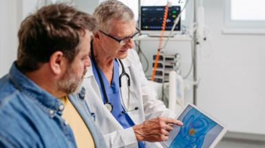 A doctor wearing a white coat and a stethoscope around his neck looks at a tablet showing the location of the prostate, with a male patient next to him. The  patient is wearing a denim shirt and has short, brown hair and a greying beard.