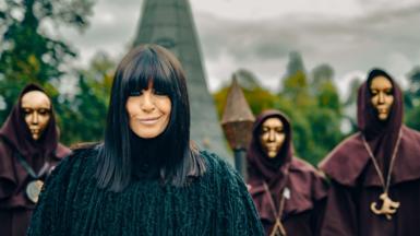 Claudia Winkleman wears a black coat as she smiles at the camera. Behind her is part of a castle and three people in black hoods and golden masks.
