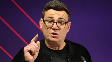 Andy Burnham making a speech. He is wearing a dark coloured jacket and glasses.