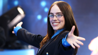 Billie Eilish smiling with her arms out stretched at the London premiere of her 3D concert film, Hit Me Hard And Soft: The Tour. She wears a blue shirt with a navy blue striped tie, and a dark coloured blazer. Her long brown hair sits on her shoulders, and she wears glasses. 