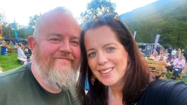 Paul McGinness has a grey beard and a shaved/balding head. He is wearing a green T-shirt. His wife Fiona has shoulder-length brown hair and sunglasses on top of her head. She is wearing a dark blue top. They are both smiling. They are at some kind of outdoor event with people in the background in a field with a hill behind. 