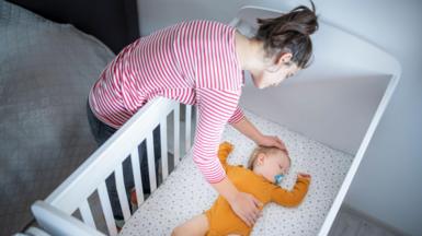 A woman leaning over a white cot, adjusting a baby lying on their back on a light-patterned mattress. The adult is wearing a red-and-white striped long-sleeved top, and the baby is dressed in an orange outfit. The scene is indoors, with a bed visible beside the cot.