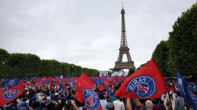 GV image showing PSG fans waving flags with the club's logo on, with the Eiffel Tower in the background