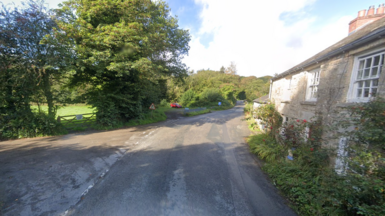 A google street view of a crossroads. On the right there is a stone house and on the left a gated green field, with tall trees. 