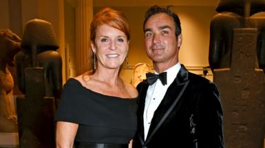 Sarah Ferguson in black dress with her hair up stands next to Manuel Fernandez who wears a black velvet jacket, white shirt and bow tie. They are both smiling at the camera. 