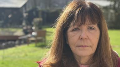 A woman with shoulder-length brown hair looks at the camera with a serious expression on her face. She is standing in a garden on a sunny day. Garden furniture and grass can be seen behind her. She is wearing a burgundy coat and pink sweater.