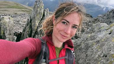 Person taking a selfie on a rocky mountain ridge, wearing a red hiking top and backpack, with rugged stone formations, grassy slopes, and dramatic cloudy skies in the background.
