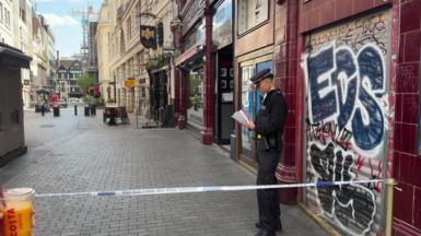 A police officer stands behind police tape on a city street in central London, reading notes outside a closed shop with graffiti on its shutter, while nearby streets are quiet and cordoned off.