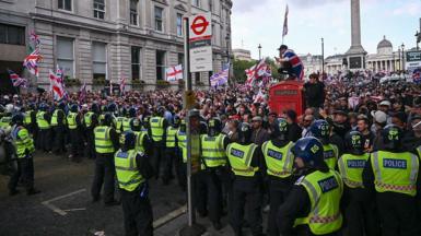 A line of police in high-visibility jackets and helmets faces a crowd waving Union Jacks and St George's Cross flags. A person draped in a flag sits atop a red telephone box near a Whitehall bus stop, with Nelson's Column in the background.
