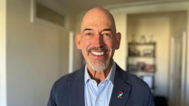 Roly Woods wearing a pale blue and white checked shirt and navy blazer, he has a special badge from the Navy which has a Pride flag on it. He is bald and has a beard. He is smiling at the camera. The background is blurred, but appears to be his home, with a shelving unit covered in items visible but blurred behind him