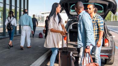 Stock photo shows a group of people removing luggage from the boot of a car as it pulls up to an airport with other passengers walking past.