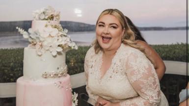 Kayley is wearing a white dress at her wedding and is stood next to a tiered cake, laughing