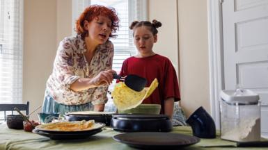 A woman with red curly hair flips a pancake on a table-top pan, using a spatula, while a younger girl, with her hair in two buns, looks on - cooked pancakes are on a plate on the table 