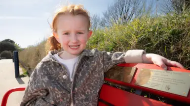 A little girl with red hair, wearing a white t shirt and leopard jacket, sat on a red bench with a gold plaque on it. Behind her is a green bush and a path.