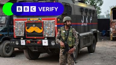 An Indian soldier stands guard. He is armed with a rifle and is wearing army fatigues. In the background is a large truck.
