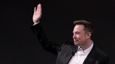 Musk in a white shirt and black jacket with his hand raised