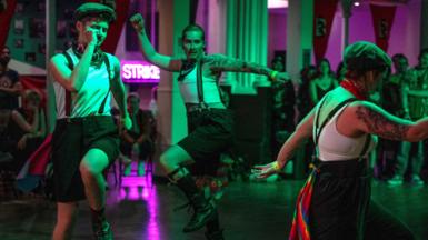 Four dancers dressed in white tops and black shorts or trousers with braces dance in a circle inside a dimly lit room.