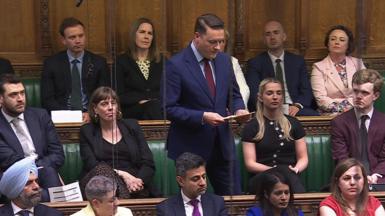 He is standing in the Commons chamber and wears a a pale blue shirt and maroon tie under a buttoned-up navy suit. He is reading from cards and is surrounded by Labour MPs who support his leadership bid.