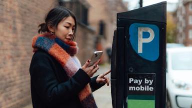Young woman holding smartphone, making mobile payment at the parking payment machine.