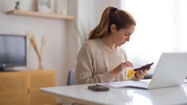 Woman sits at a desk in a home with a phone and a laptop in front of her and a calculator in her hand.