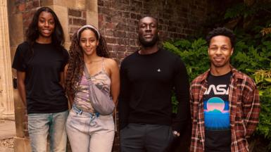The rapper Stormzy with student Destiny, Drew and Dimario alongside him. They stand outside a brick building with shrubbery.