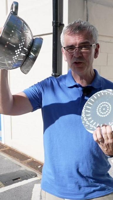 A man holding up a colander and a white paper plate, which has shadows cast on it.