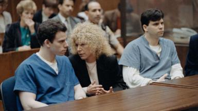 The Menendez brothers, wearing different shades of a blue prison uniform, sit at a table with one of their attorneys during their trial in 1994. Inside the courtroom, their attorney is a woman with curly hair and appears deep in conversation with Erik Menendez. 