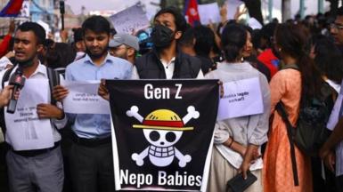 A man in a black facemask holds a poster saying Gen Z Nepo Babies, with a skull and crossbones in the middle. Around him other protesters stand, holding printed signs. Flags can be seen being waved in the background