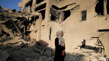 A woman wearing a white flowered hijab and black dress stands in rubble of a home in Gaza 